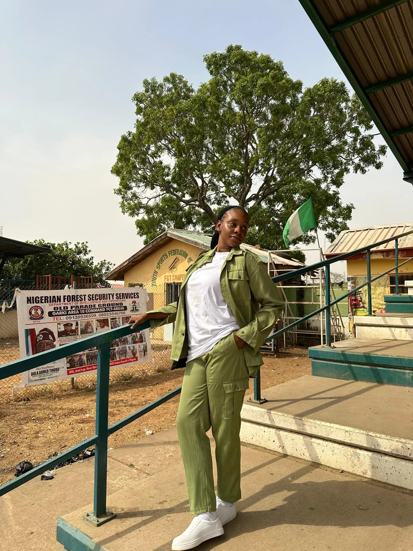 Peace in her NYSC uniform at camp, smiling beside the Nigerian flag